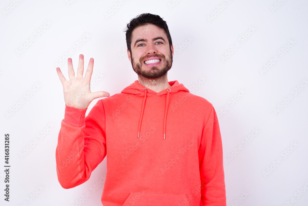 young Caucasian bearded man wearing pink hoodie against white wall showing and pointing up with fingers number five while smiling confident and happy. young Caucasian bearded man wearing pink hoodie against white wall showing and pointing up with fingers number five while smiling confident and happy.