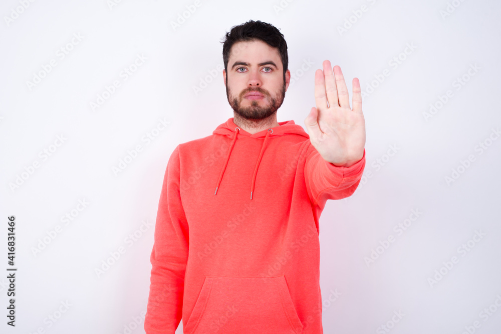 young Caucasian bearded man wearing pink hoodie against white wall doing stop gesture with palm of the hand. Warning expression with negative and serious gesture on the face. young Caucasian bearded man wearing pink hoodie against white wall doing stop gesture with palm of the hand. Warning expression with negative and serious gesture on the face.
