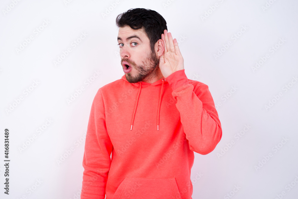 Oh my God! Funny astonished young Caucasian bearded man wearing pink hoodie against white wall opening mouth widely looking aside, with hand near ear trying to listen to gossips. Oh my God! Funny astonished young Caucasian bearded man wearing pink hoodie against white wall opening mouth widely looking aside, with hand near ear trying to listen to gossips.