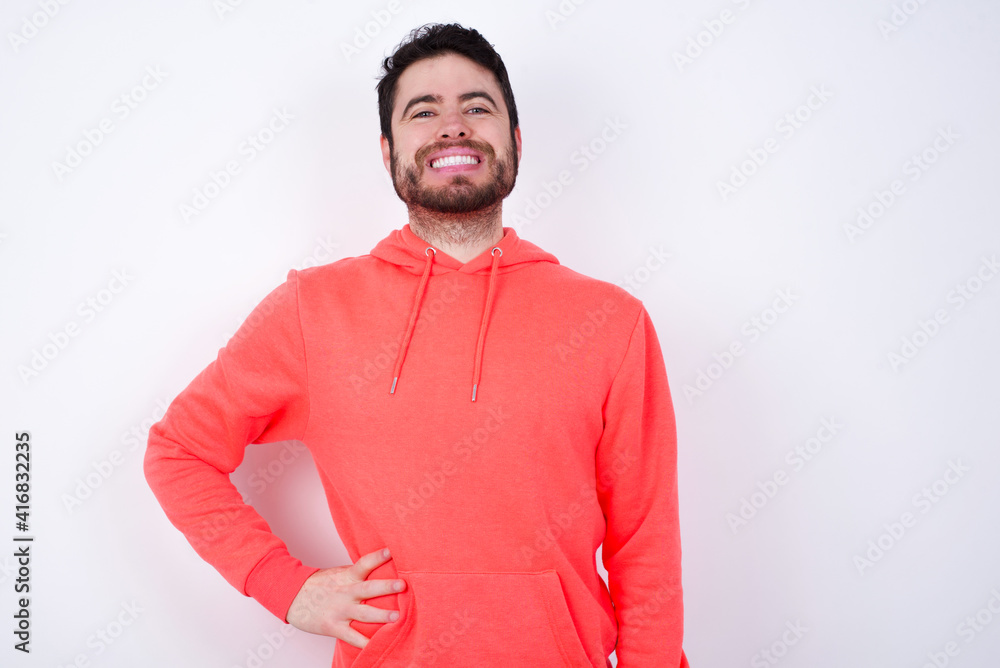 Fototapeta premium Studio shot of cheerful young Caucasian bearded man wearing pink hoodie against white wall keeps hand on hip, smiles broadly.
