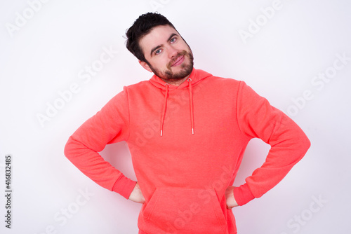 Funny frustrated young Caucasian bearded man wearing pink hoodie against white wall holding hands on waist and silly looking at awkward situation.