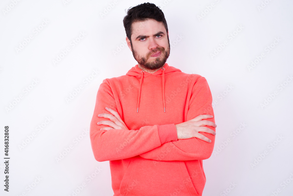 Picture of angry young Caucasian bearded man wearing pink hoodie against white background crossing arms. Looking at camera with disappointed expression.