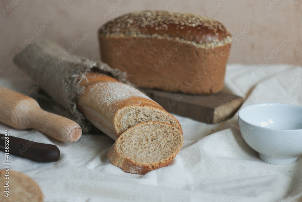 On the table, a piece of baguette was cut from a loaf, and in the background there is bread with grains on textiles
