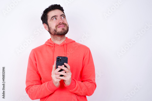 Happy young Caucasian bearded man wearing pink hoodie against white background listening to music with earphones using mobile phone. © Jihan