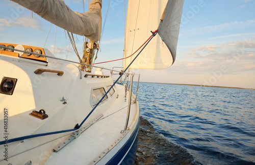 Blue sloop rigged yacht sailing in the sea at sunset. Clear sky. A view from the deck to the bow, mast, sails. Transportation, travel, cruise, sport, recreation, leisure activity, racing, regatta photo