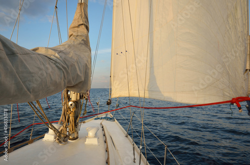 White sloop rigged yacht sailing in the sea at sunset. Clear sky. A view from the deck to the bow, mast, sails. Transportation, travel, cruise, sport, recreation, leisure activity, racing, regatta photo