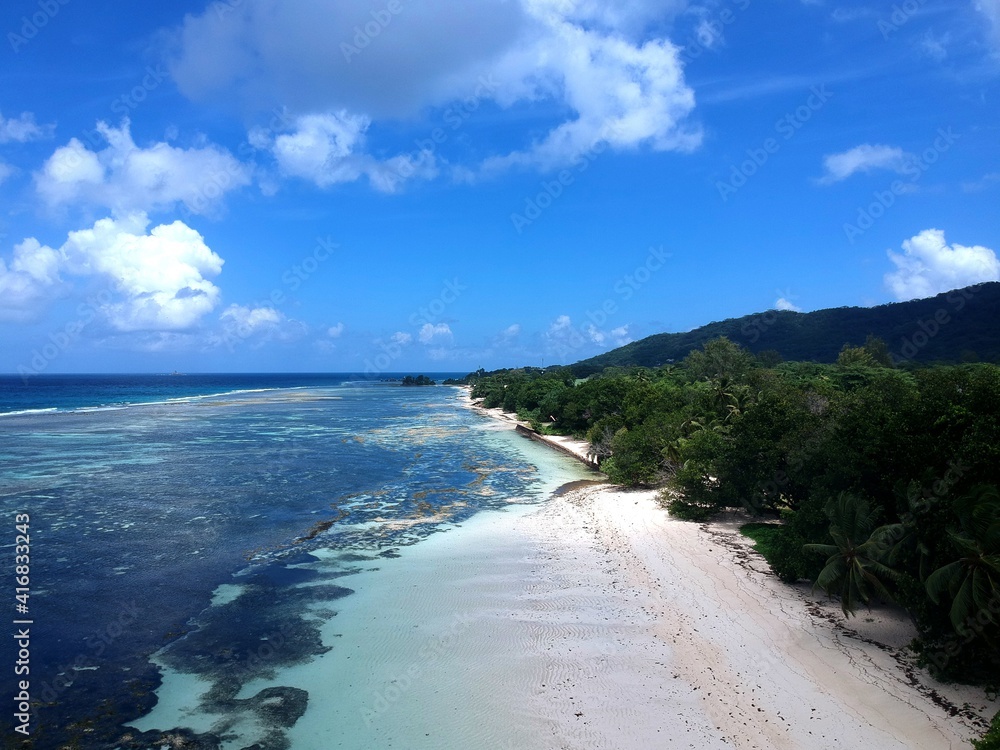 aerial view of Seychelles La Digue Island coastline with white sandy beach, green trees, blue turquoise clear water, reef and blue sky with white clouds