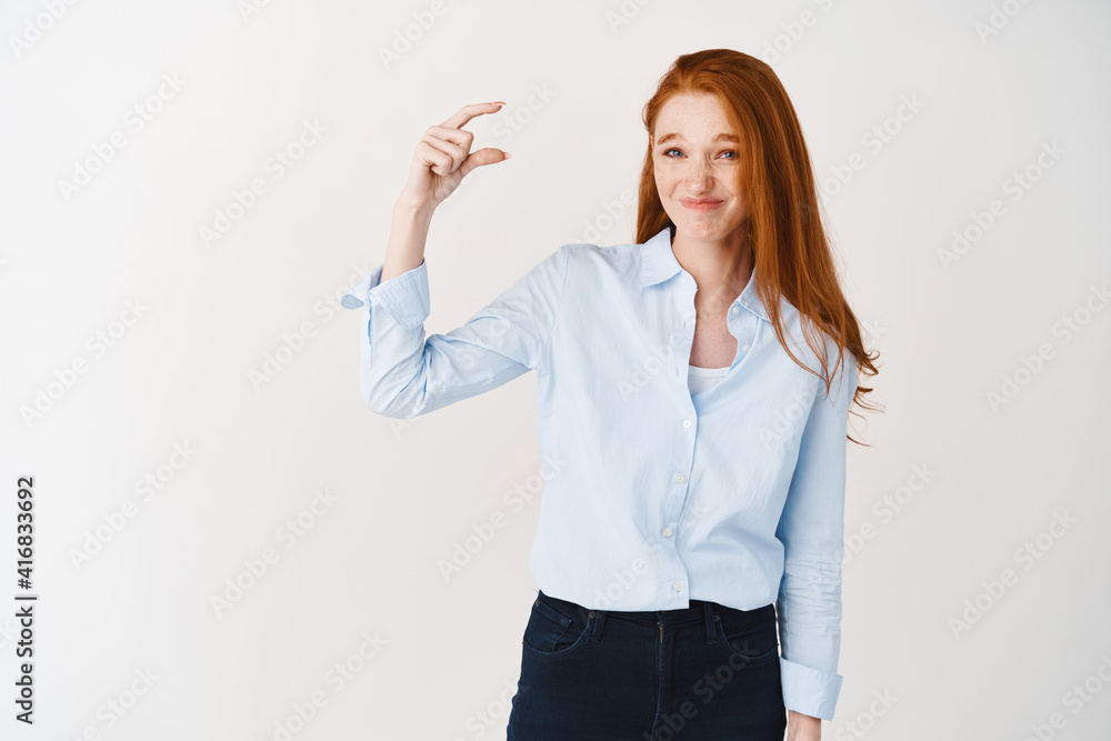 Disappointed redhead girl in blue shirt showing something small and grimacing awkward, judging something tiny or little, standing over white background Disappointed redhead girl in blue shirt showing something small and grimacing awkward, judging something tiny or little, standing over white background