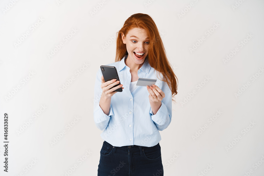 Cheerful girl shopping online, making purchase in smartphone app with plastic credit card, standing against white background Cheerful girl shopping online, making purchase in smartphone app with plastic credit card, standing against white background