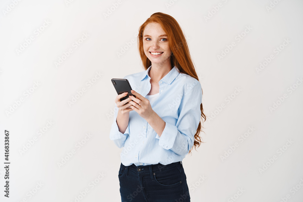 Happy pretty lady with ginger hair and blue eyes using mobile phone, smiling at camera, standing against white background Happy pretty lady with ginger hair and blue eyes using mobile phone, smiling at camera, standing against white background