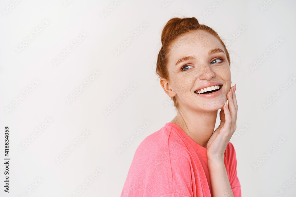 Beauty and skincare. Smiling girl with ginger hair combed in messy bun, touching perfect and glowing skin and smiling, showing face without makeup, white background Beauty and skincare. Smiling girl with ginger hair combed in messy bun, touching perfect and glowing skin and smiling, showing face without makeup, white background