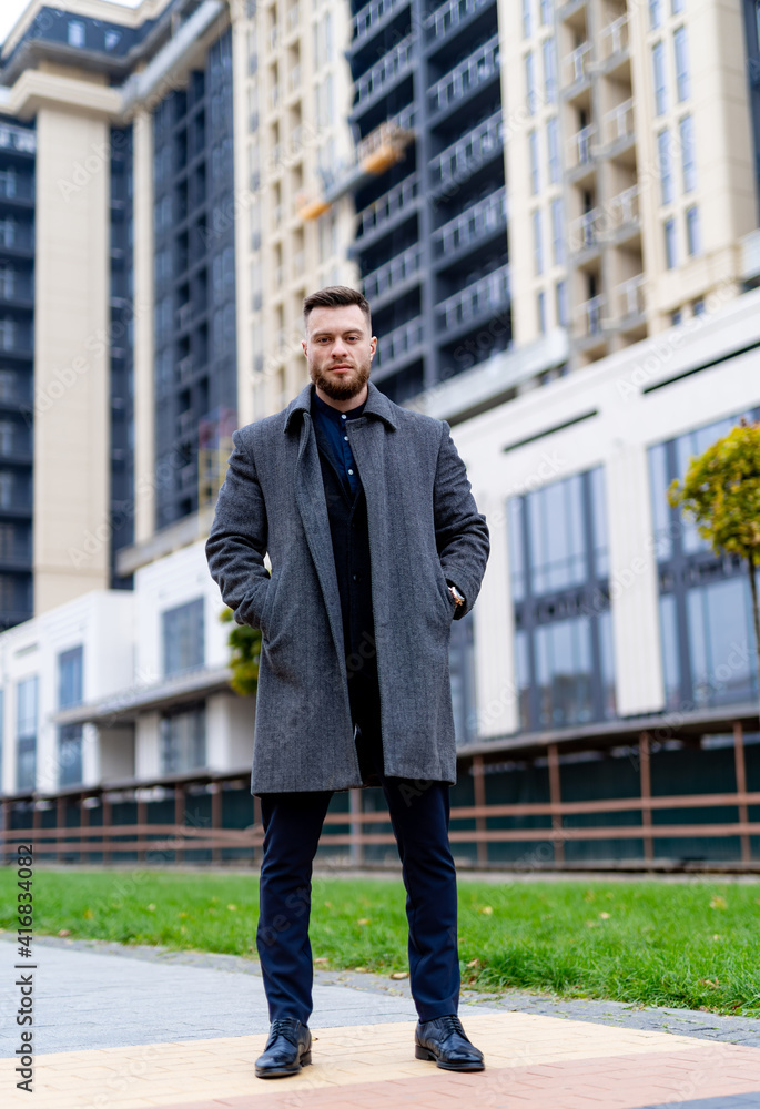 Man in gray coat and black shirt on background of business skyscrapers, looking at the camera. Businessman in coat standing near international business centre. Man in gray coat and black shirt on background of business skyscrapers, looking at the camera. Businessman in coat standing near international business centre.