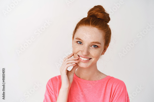 Beauty and skincare. Close-up of cute teenage girl with ginger hair combed in bun, smiling with white teeth, showing glowing skin, no makeup on face, standing over white background