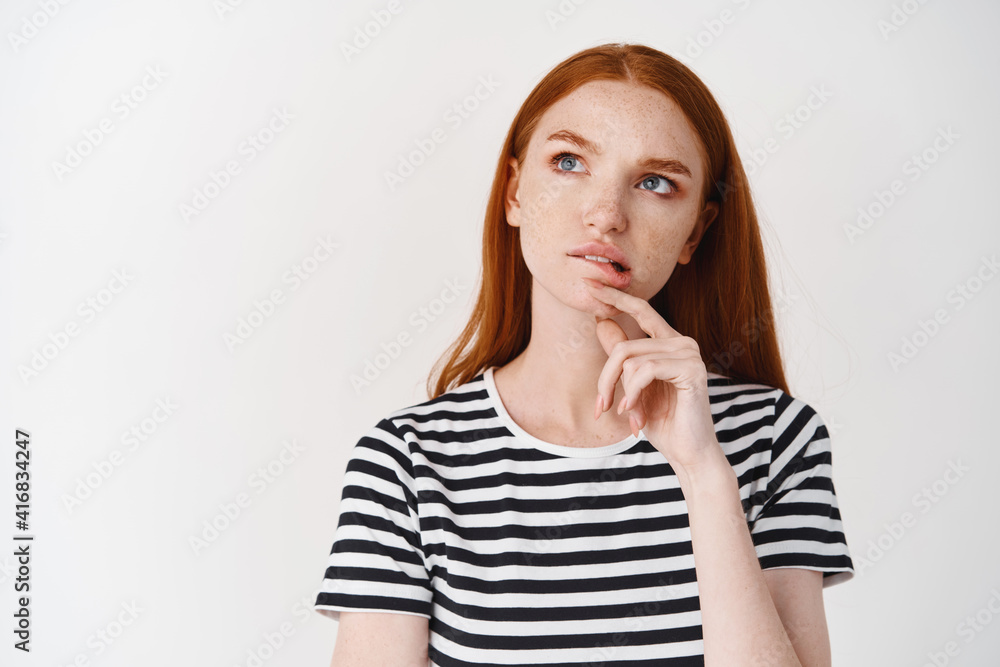Thoughtful teen girl with freckles and red hair biting lip, frowning and looking up while thinking, making choice, standing over white background Thoughtful teen girl with freckles and red hair biting lip, frowning and looking up while thinking, making choice, standing over white background