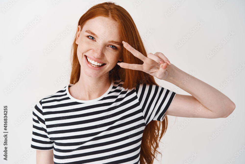 Beautiful young woman with long ginger hair, showing peace sign and smiling happy, standing over white background Beautiful young woman with long ginger hair, showing peace sign and smiling happy, standing over white background
