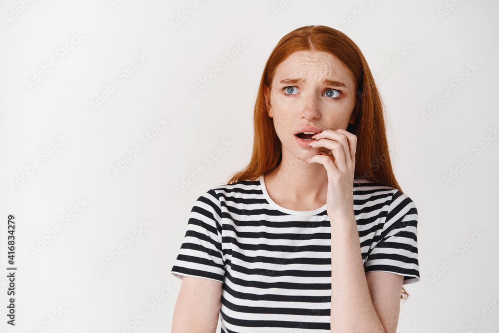 Anxious redhead girl biting finger and looking doubtful left, frowning worried, standing over white background Anxious redhead girl biting finger and looking doubtful left, frowning worried, standing over white background