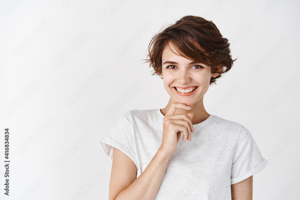 Portrait of candid happy woman smiling and touching chin, looking pensive, standing against white background Portrait of candid happy woman smiling and touching chin, looking pensive, standing against white background