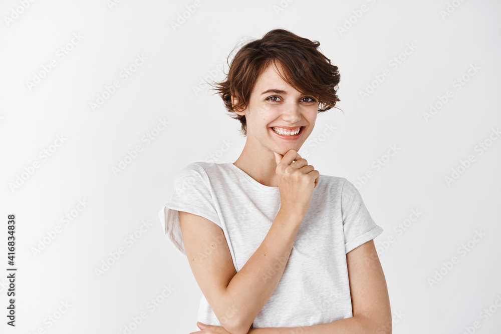 Portrait of pensive european woman with short hair, touching chin and smiling thoughtful, looking at camera, standing in t-shirt on white background Portrait of pensive european woman with short hair, touching chin and smiling thoughtful, looking at camera, standing in t-shirt on white background