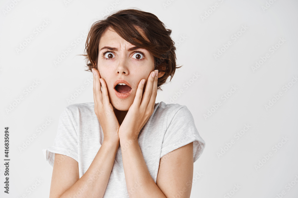 Shocked and startled caucasian woman gasping and frowning, looking at something terrible, standing on white background in casual t-shirt Shocked and startled caucasian woman gasping and frowning, looking at something terrible, standing on white background in casual t-shirt