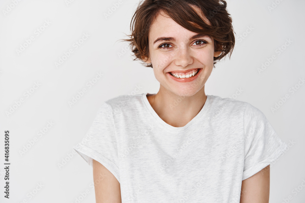 Close-up of beautiful and tender woman with short hair, smiling and looking cute at camera, wearing t-shirt, standing against white background Close-up of beautiful and tender woman with short hair, smiling and looking cute at camera, wearing t-shirt, standing against white background
