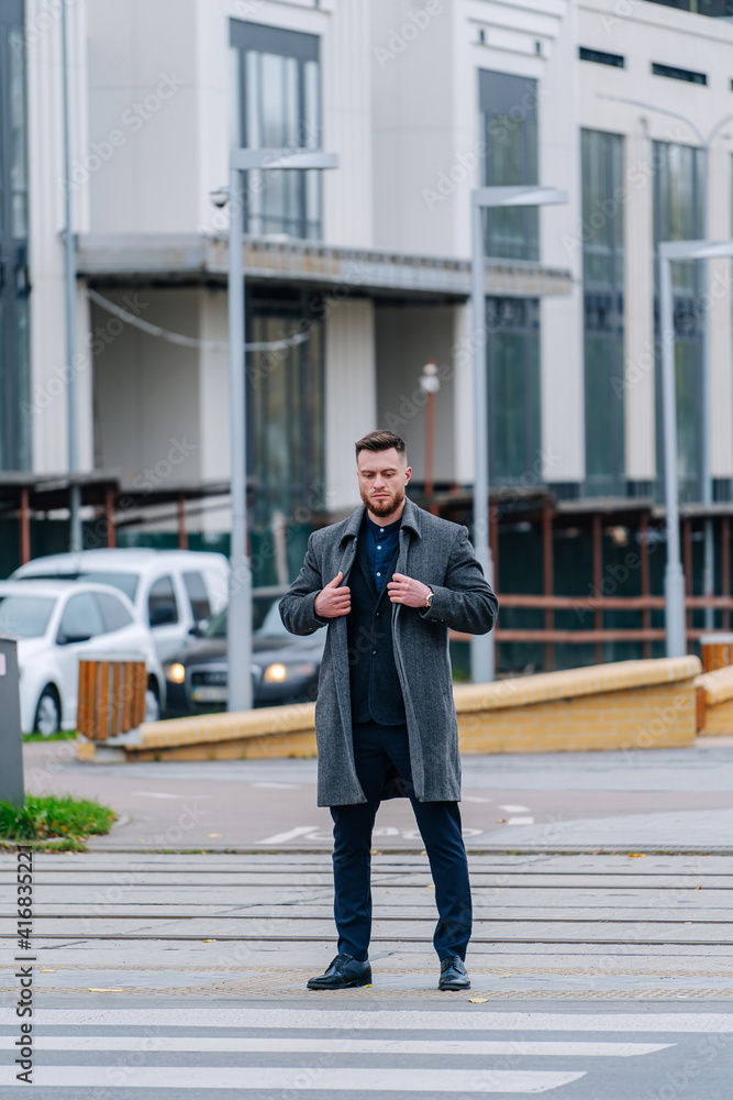 Stylish male in formal clothes. Model man wears suit and gray coat. Modern building with good architecture on the background.