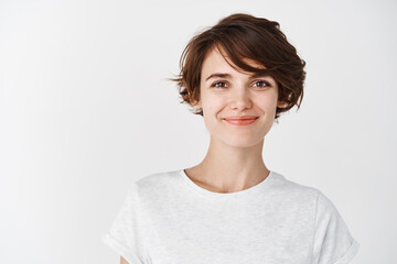Head shot of beautiful caucasian woman with short haircut, smiling and looking confident, standing in t-shirt on white background
