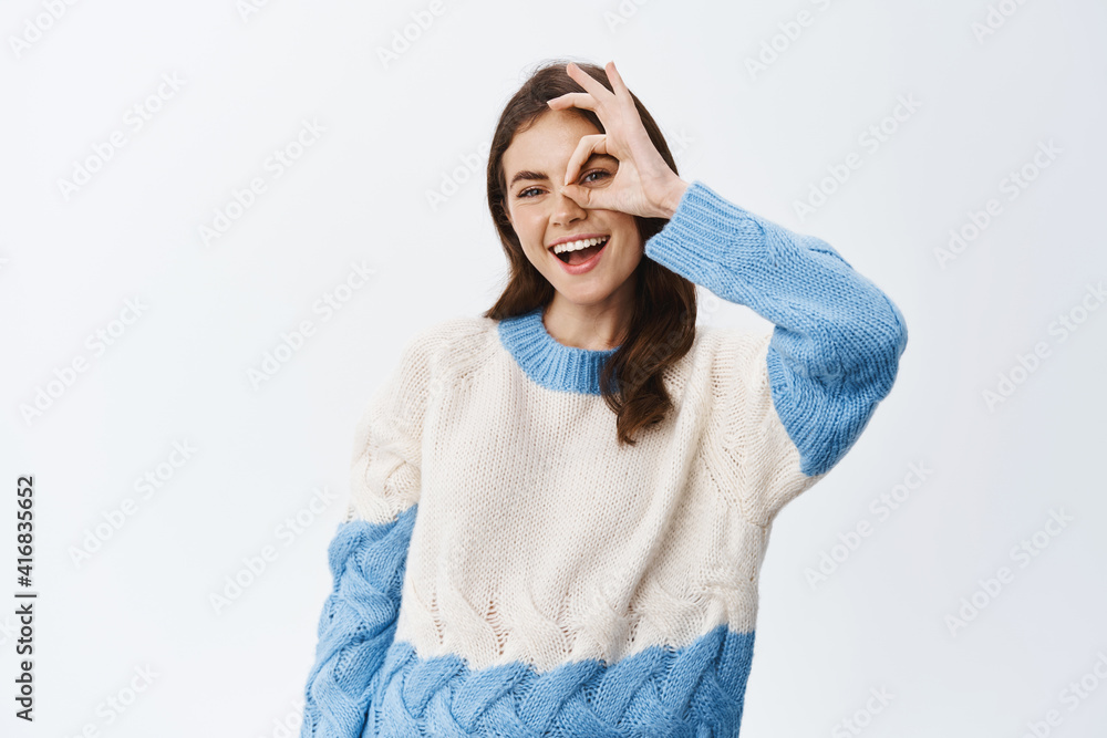 Excited smiling woman tell yes, showing okay sign over eye, looking through OK gesture with happy pleased face, standing in sweater against white background Excited smiling woman tell yes, showing okay sign over eye, looking through OK gesture with happy pleased face, standing in sweater against white background