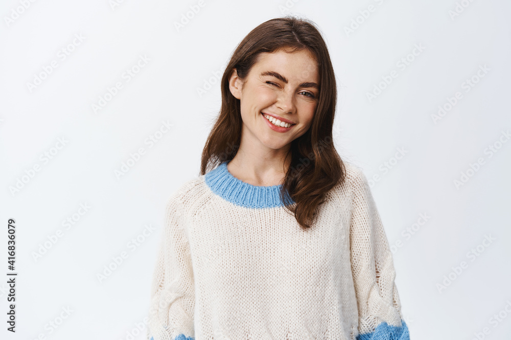 Portrait of young confident woman in sweater, smiling and winking assuringly, standing happy against white background Portrait of young confident woman in sweater, smiling and winking assuringly, standing happy against white background