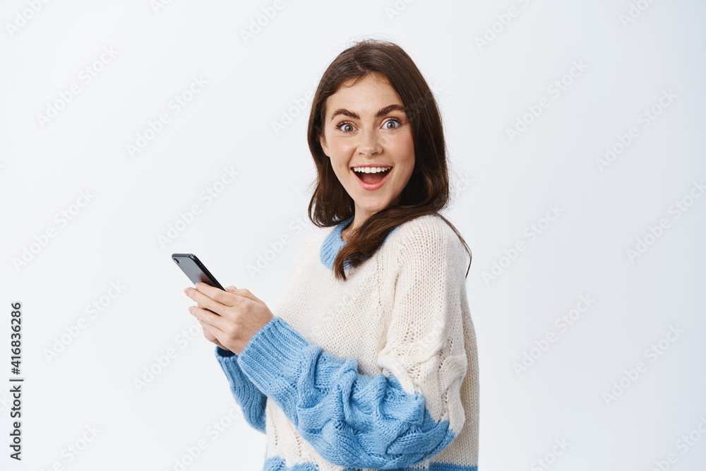 Portrait of cheerful brunette woman turn head at camera after reading news on mobile phone screen, smiling excited, using smartphone application, white background Portrait of cheerful brunette woman turn head at camera after reading news on mobile phone screen, smiling excited, using smartphone application, white background