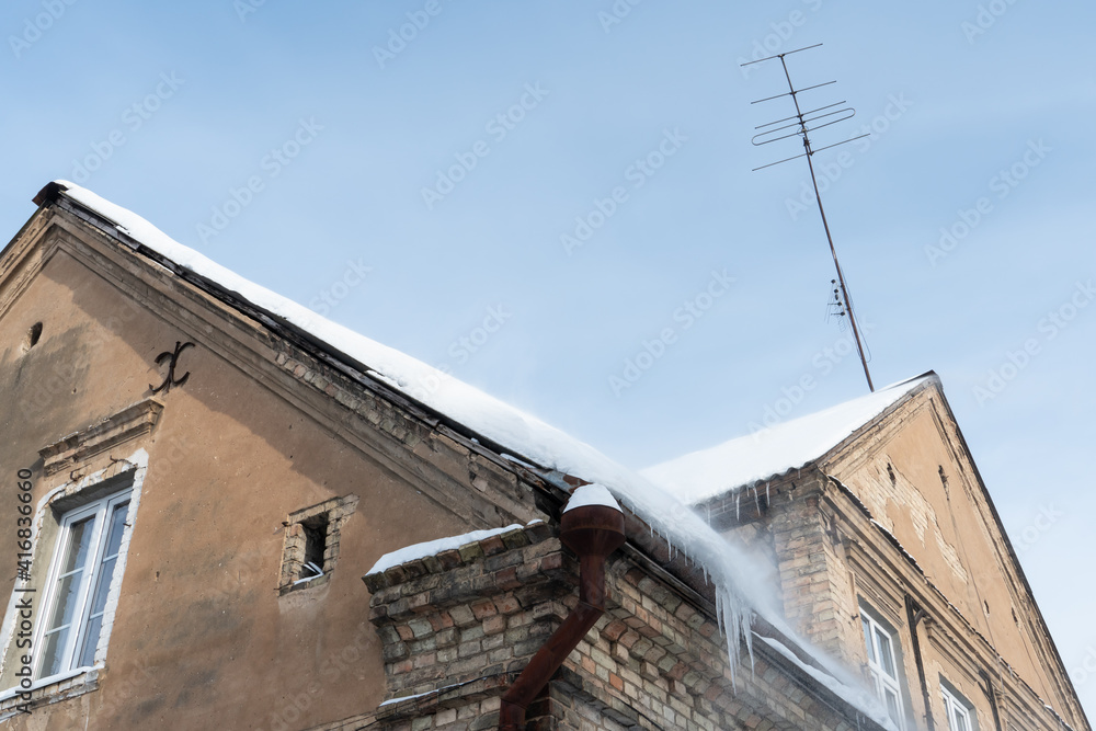 Old brick building covered in snow of heavy snowfall. Hanging icicles from the roof of a residential building. View of the red brick building against the blue sky. danger from falling icicles