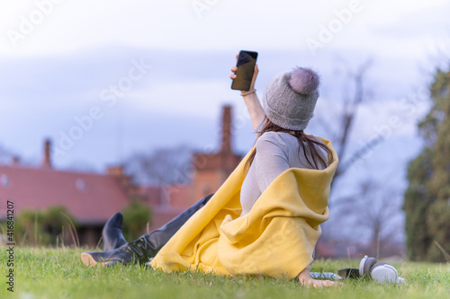 Fototapeta Naklejka Na Ścianę i Meble -  Beautiful young woman in gray dress, yellow scarf and hat sitting in a park on the grass taking a smartphone selfie after work at sunset in spring. relaxation lifestyle concept. businesswoman