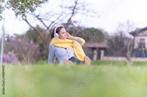 beautiful young woman in gray dress, yellow scarf and hat sitting in a park on the grass hearing music wearing headphones. relaxation lifestyle concept. businesswoman