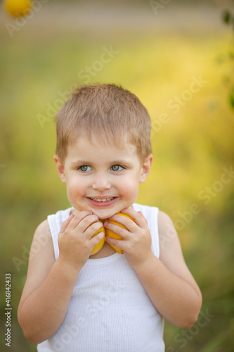 A cute boy with blond hair in a white T-shirt with summer lemons in the garden under a tree