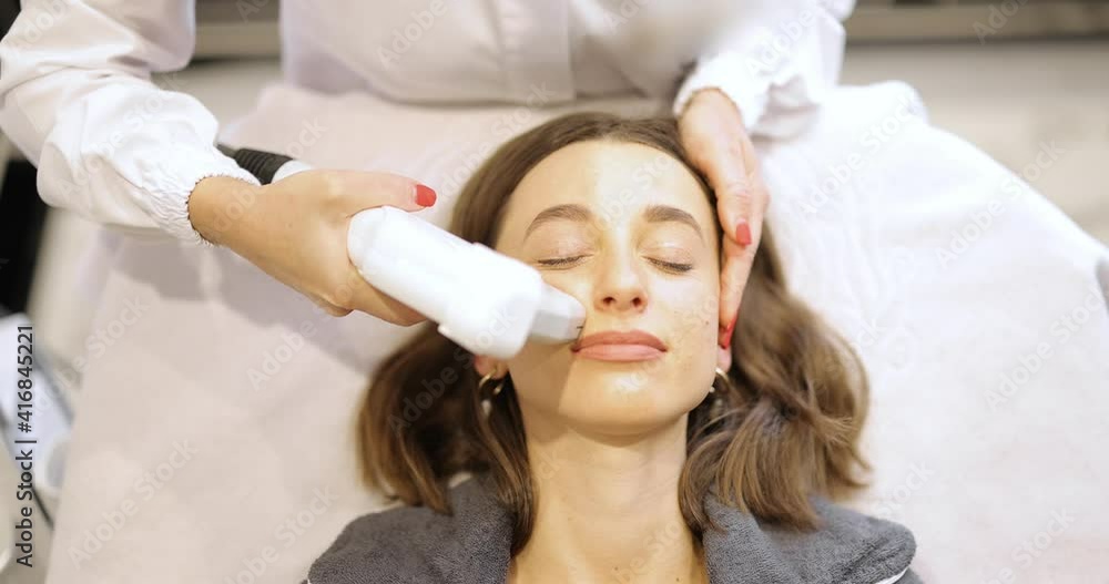 Young adult woman during an ultrasound face lifting procedure in beauty clinic with cosmetologist. Anti-age and wellness concept Young adult woman during an ultrasound face lifting procedure in beauty clinic with cosmetologist. Anti-age and wellness concept