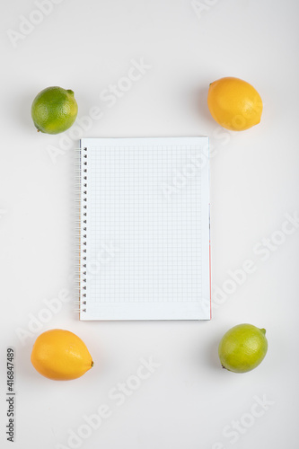 Ripe juicy lemons and limes around white notebook over white wall