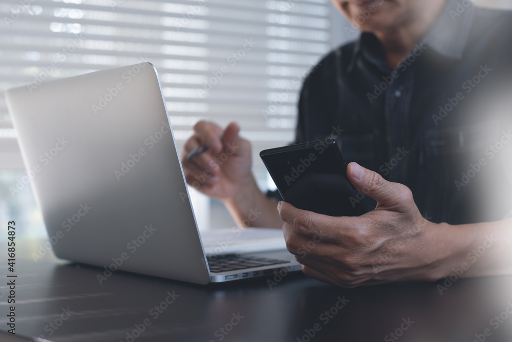 Man using mobile smartphone during working on laptop computer from home office, close up Man using mobile smartphone during working on laptop computer from home office, close up