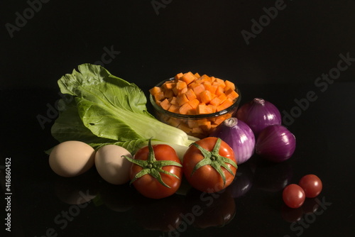 Various fresh ingredients isolated on black background with a mirror reflection. A bowl of diced carrots. Bok choy, tomatoes, cherry tomatoes, purple onions and eggs