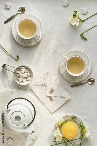 Springtime afternoon tea. Spring freesia flowers, yellow candle, white teapot and tea cup on table. Flat lay, off white textile tablecloth. Easter eggs, white ceramic cups and sugar hearts.