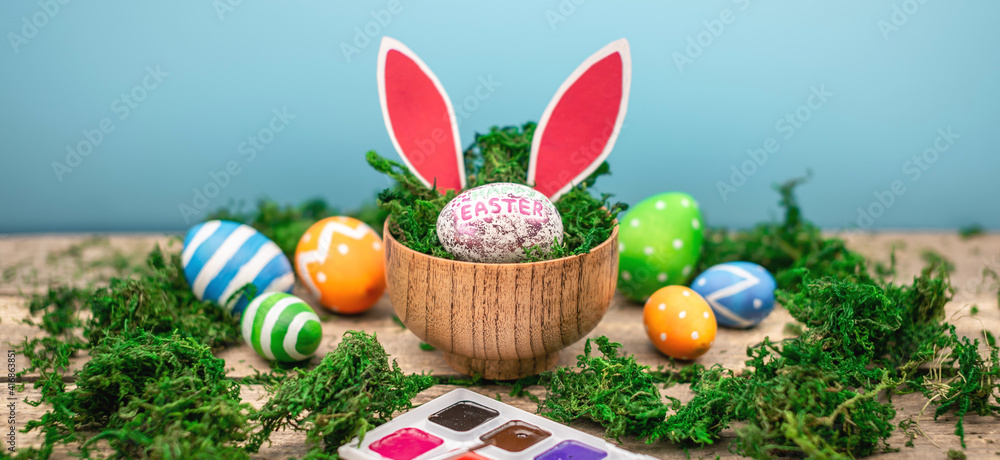 Bright colorful Easter eggs and rabbit ears on a blue and wooden background with green moss Bright colorful Easter eggs and rabbit ears on a blue and wooden background with green moss
