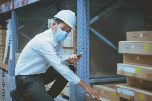Asian handsome engineer or technician wearing mask,and safety hard hat,uses digital tablet check merchandise stock,security cargo management,in warehouse,industry business logistic and export concept