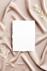 Blank white paper card on satin fabric with dried flowers. Flat lay, top view, copy space. Wedding invitation card design.
