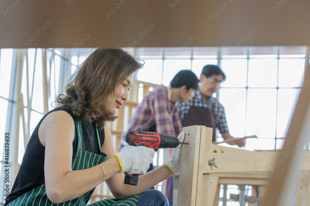 asian adult female worker wearing black shirt with green stripe apron and white gloves sit drilling wooden stick with red electric screwdriver when her male colleagues wearing plaid shirt discussing asian adult female worker wearing black shirt with green stripe apron and white gloves sit drilling wooden stick with red electric screwdriver when her male colleagues wearing plaid shirt discussing