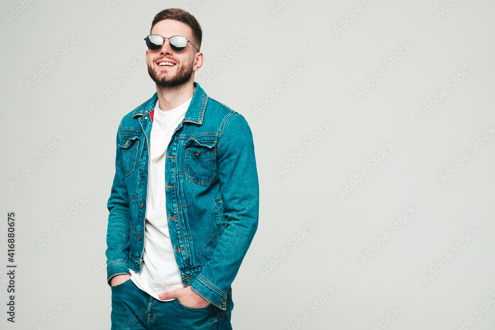 Portrait of handsome smiling stylish hipster lambersexual model.Man dressed in jacket and jeans clothes. Fashion male posing on grey background in studio un sunglasses