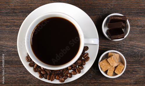 White porcelain cup of coffee  saucer with coffee beans  bowls with cane brown sugar and dark chocolate on dark wooden background