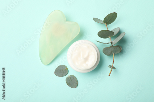 Jade gua sha tool, cream and eucalyptus branch on light blue background, flat lay photo