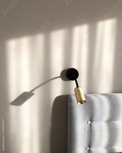 Minimalism and simplicity in bedroom decor. Beautiful light shining on wall with gold sconce and gray bed headrest. Scandinavian style.