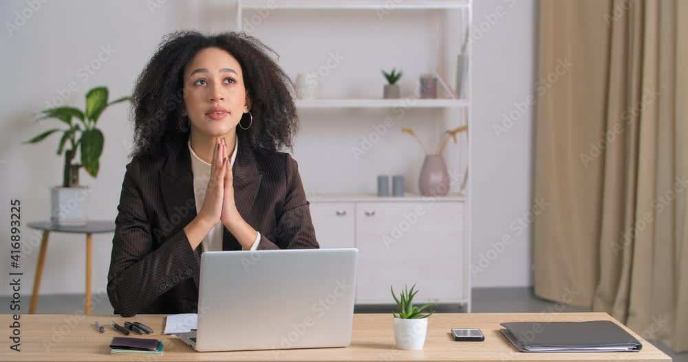 Afro american mixed race business woman student sitting at table with laptop feeling tired folds hands palms in supplication pray request hope for luck help destiny good results promotion at work Afro american mixed race business woman student sitting at table with laptop feeling tired folds hands palms in supplication pray request hope for luck help destiny good results promotion at work