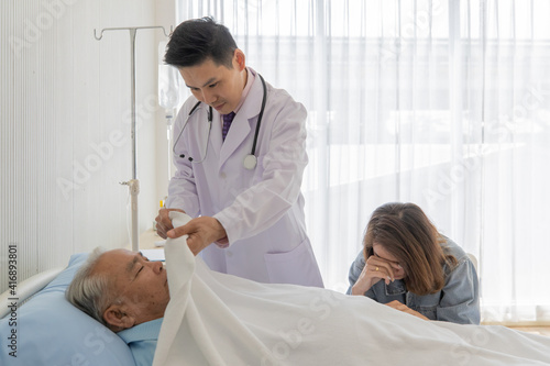 A young asian male doctor wearing white lab coat and stethoscope standing next to patient bed use white blanket covering patient dead body on the hospital bed while female cousin crying nearby