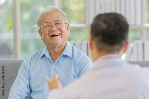 An old fat male patient with gray white hair wearing light blue shirt and sitting on sofa laughing and discussing about health with a young doctor with black hair wearing white lab coat