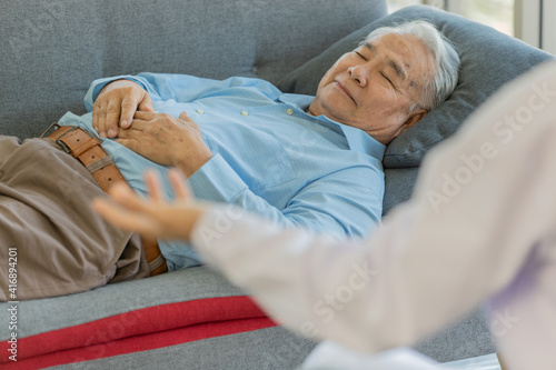 Doctor wearing white lab coat open his left hand up when explaining to old fat asian male patient wearing light blue shirt laying down holding his hand together on gray sofa in blurred background